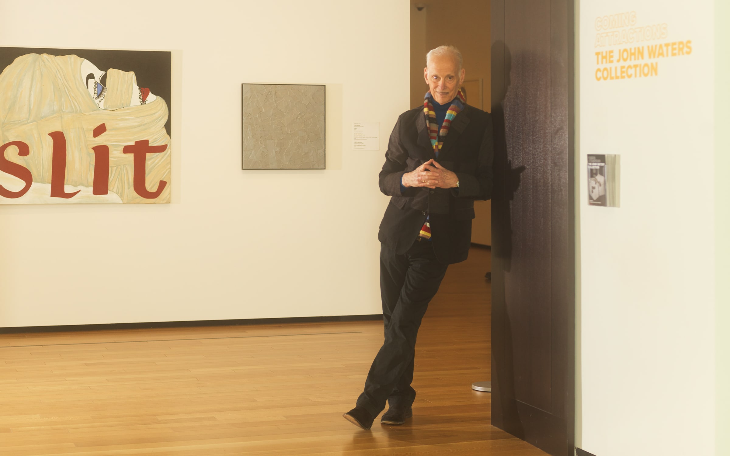 John Waters at the entrance of 'Coming Attractions', the exhibition of his collection at the Baltimore Museum of Art, January 2023. In the background are Kathe Burkhart's Slit: from the Liz Taylor Series (Ash Wednesday) (1992) and Tom Sachs's What (1995). Photograph by Matt Grubb for Art Basel.